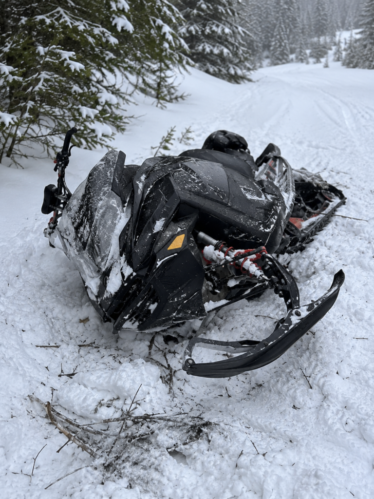 Damaged snowmobile after a winter accident on a snowy forest trail.